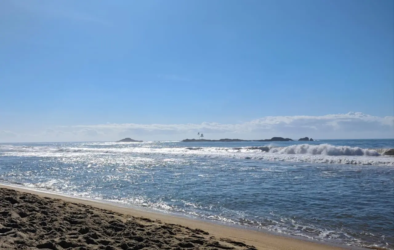 The shore at Vatomandry: wet sand in the foreground, waves breaking, and low rocky islets on the horizon under a clear sky.