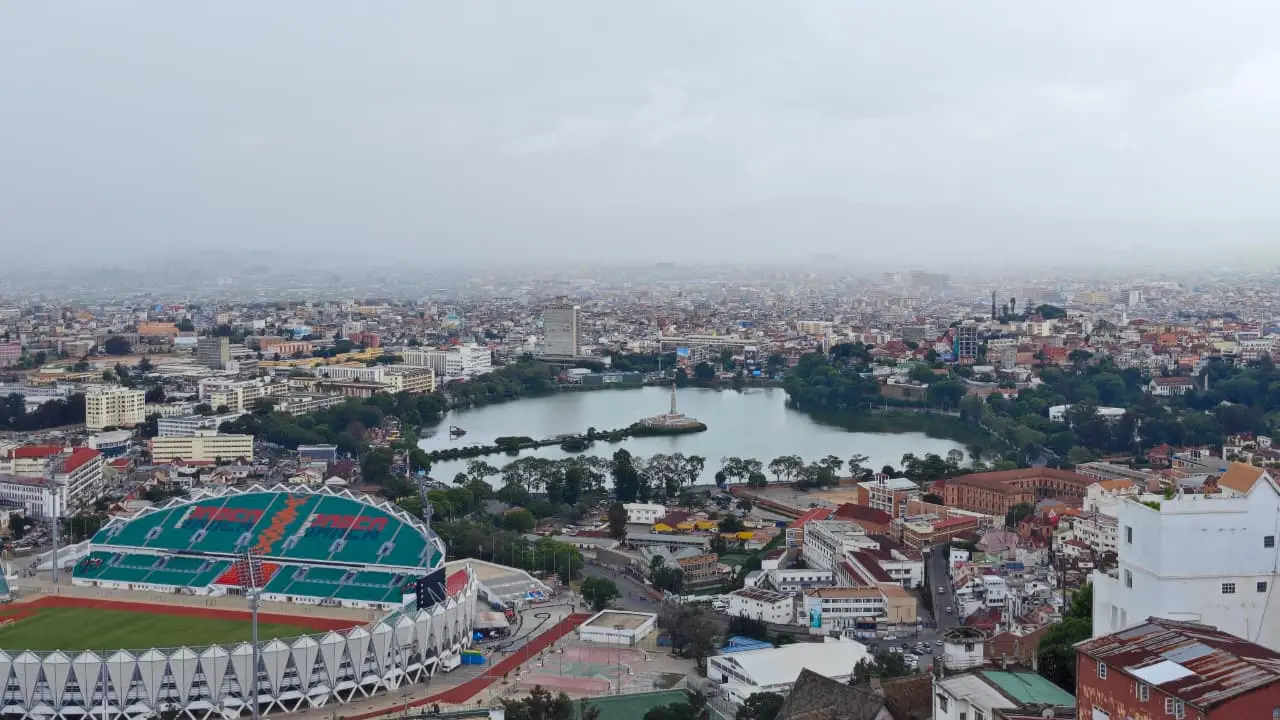 Antananarivo seen from above: a lake with a monument at its centre, a stadium at lower left, and dense housing spreading to the hills under an overcast sky.