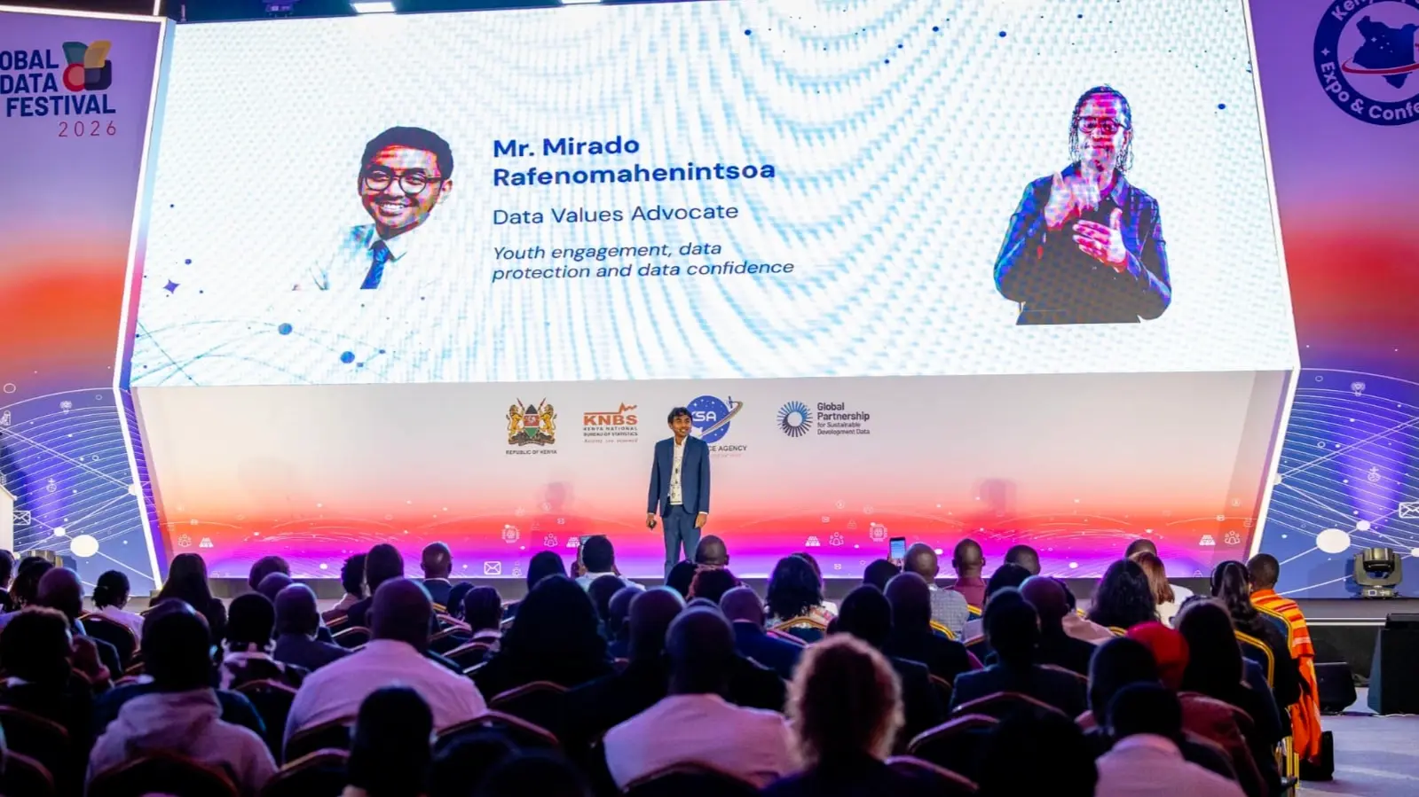 Speaking on stage at the Global Data Festival in Nairobi, facing a seated audience, with a large screen behind showing the session title and a sign-language interpreter.
