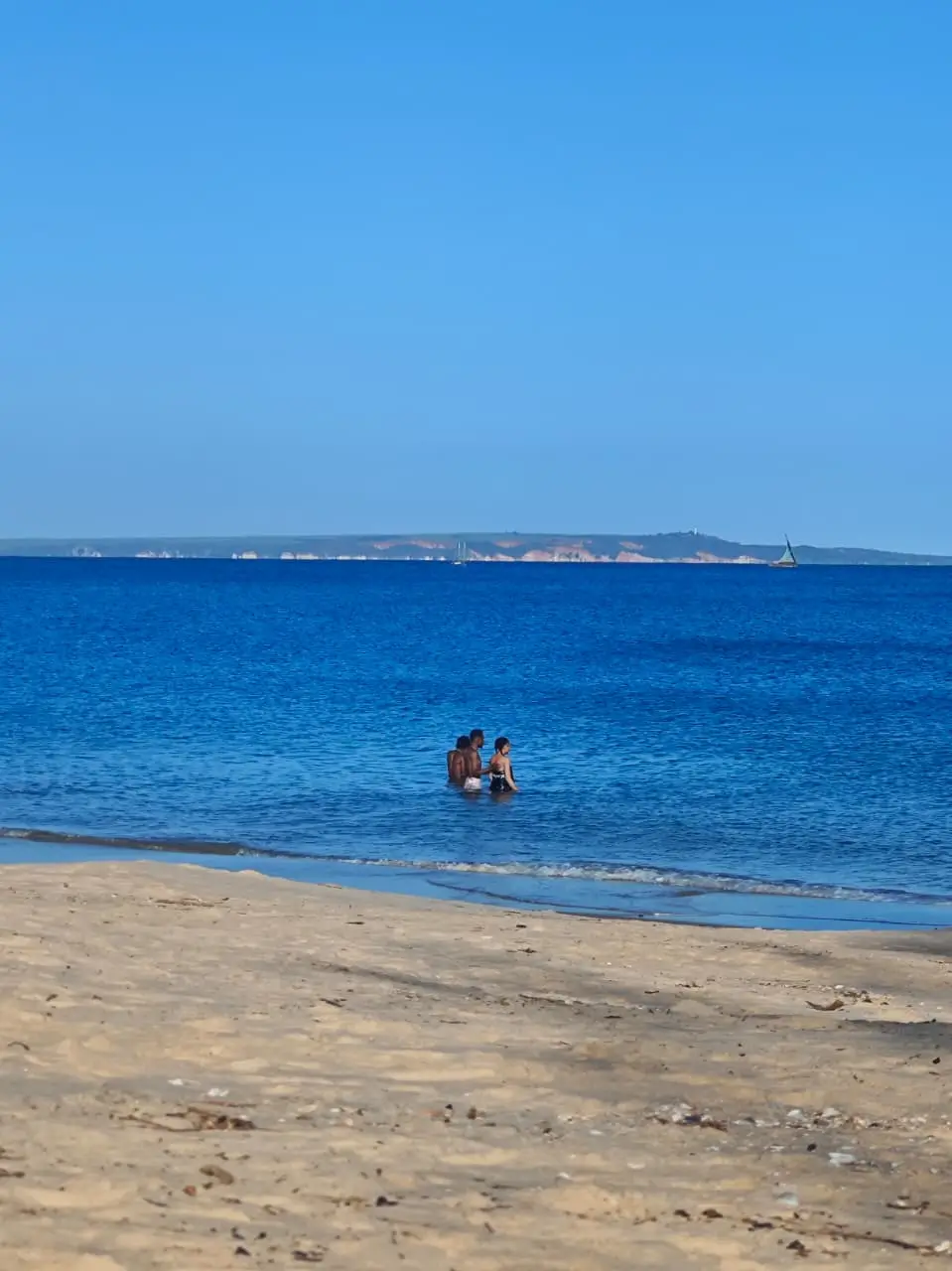 The beach at Mahajanga: pale sand, two people standing in shallow blue water, and a low headland with a sailing boat on the horizon.